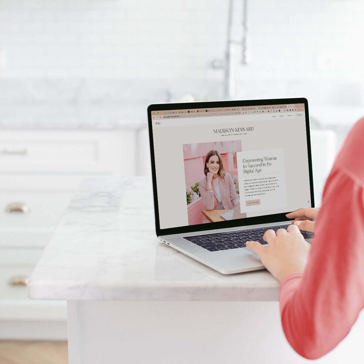 A woman working on a laptop at a marble countertop, with a web page titled “Empowering Women to Succeed in the Digital Age” displayed on the screen.